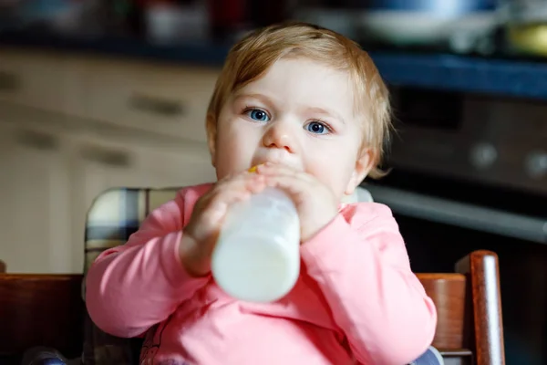 Roztomilá roztomilá holčička držící kojící láhev a pijící mléko. První jídlo pro děti. Novorozené dítě, sedící v křesle domácí kuchyně. Zdravé děti a koncepce krmení láhví — Stock fotografie