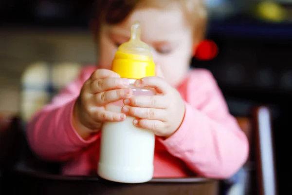 Linda menina adorável segurando mamadeira e bebendo leite fórmula. Primeira comida para bebés. Criança recém-nascida, sentada na cadeira da cozinha doméstica. Bebês saudáveis e conceito de alimentação por mamadeira — Fotografia de Stock