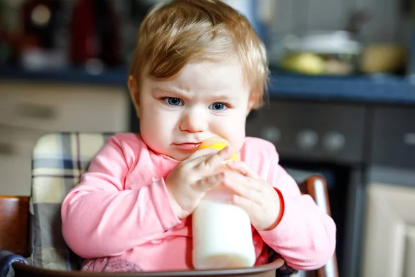 Linda niña adorable sosteniendo biberón y beber leche maternizada. Primera comida para bebés. Niño recién nacido, sentado en la silla de la cocina doméstica. Bebés sanos y concepto de alimentación con biberón — Foto de Stock