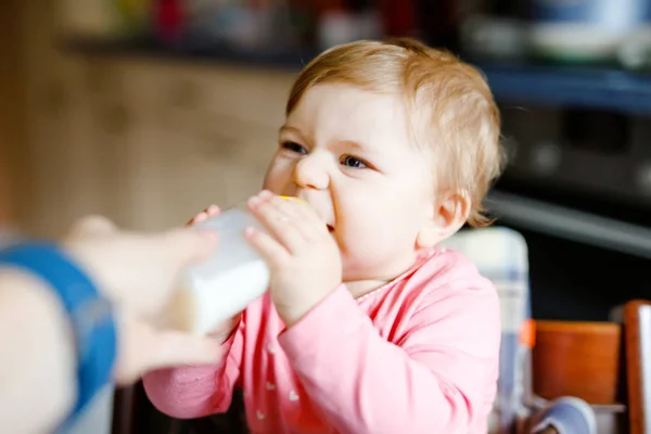 Jolie adorable petite fille tenant le biberon et le lait maternisé. Première nourriture pour bébés. Enfant nouveau-né, assis dans la chaise de la cuisine domestique. Bébés en santé et concept d'alimentation au biberon — Photo