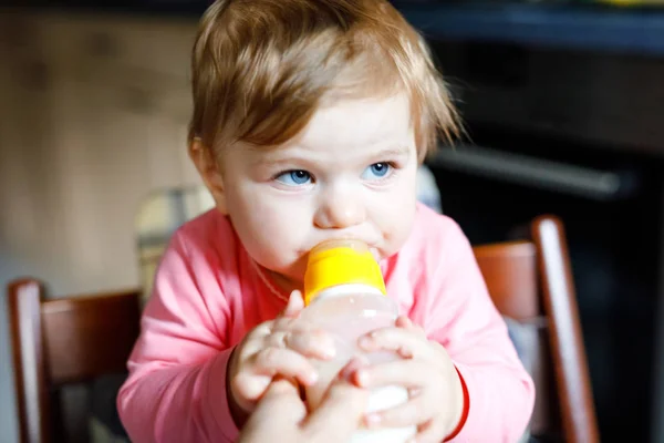 Linda menina adorável segurando mamadeira e bebendo leite fórmula. Primeira comida para bebés. Criança recém-nascida, sentada na cadeira da cozinha doméstica. Bebês saudáveis e conceito de alimentação por mamadeira — Fotografia de Stock