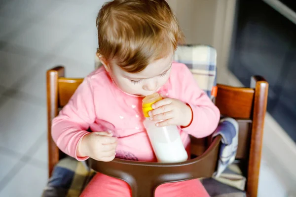 Linda menina adorável segurando mamadeira e bebendo leite fórmula. Primeira comida para bebés. Criança recém-nascida, sentada na cadeira da cozinha doméstica. Bebês saudáveis e conceito de alimentação por mamadeira — Fotografia de Stock