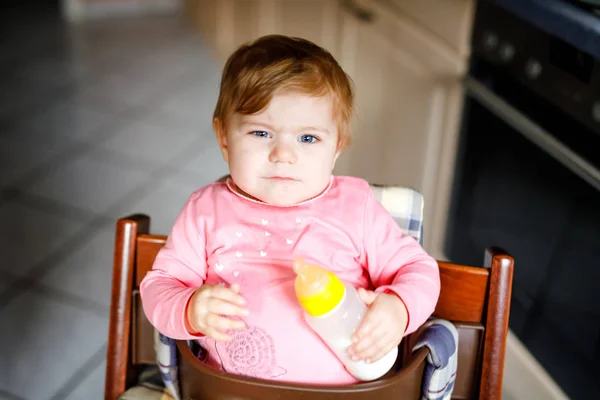 Linda niña adorable sosteniendo biberón y beber leche maternizada. Primera comida para bebés. Niño recién nacido, sentado en la silla de la cocina doméstica. Bebés sanos y concepto de alimentación con biberón — Foto de Stock