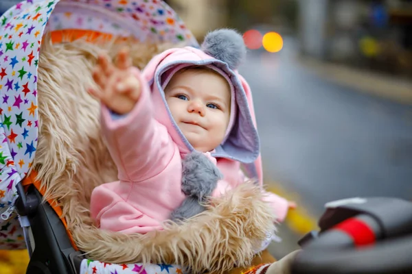 Schattig klein mooi babymeisje zittend in de kinderwagen of kinderwagen op herfstdag. Gelukkig lachend kind in warme kleren, mode stijlvolle roze babyjas met konijnenoren. Baby gaat wandelen met ouders. — Stockfoto