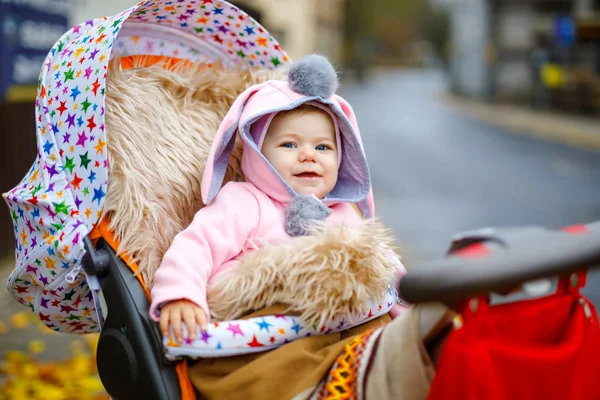 Cute little beautiful baby girl sitting in the pram or stroller on autumn day. Happy smiling child in warm clothes, fashion stylish pink baby coat with bunny ears. Baby going on a walk with parents. — Stock Photo, Image