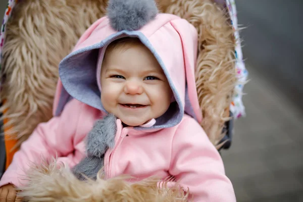 Cute little beautiful baby girl sitting in the pram or stroller on autumn day. Happy smiling child in warm clothes, fashion stylish pink baby coat with bunny ears. Baby going on a walk with parents. — Stock Photo, Image