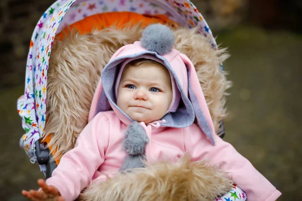 Cute little beautiful baby girl sitting in the pram or stroller on autumn day. Happy smiling child in warm clothes, fashion stylish pink baby coat with bunny ears. Baby going on a walk with parents. — Stock Photo, Image