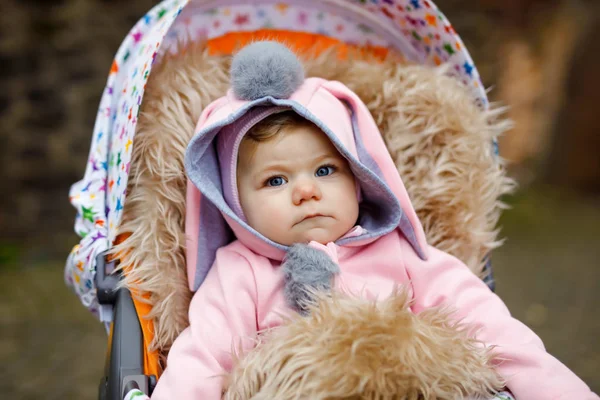 Cute little beautiful baby girl sitting in the pram or stroller on autumn day. Happy smiling child in warm clothes, fashion stylish pink baby coat with bunny ears. Baby going on a walk with parents. — Stock Photo, Image