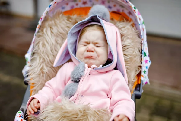 Triste llorando niña hermosa sentada en el cochecito o cochecito en el día de otoño. Niño infeliz cansado y agotado en ropa de abrigo, abrigo de bebé rosa con estilo de moda con orejas de conejo . — Foto de Stock