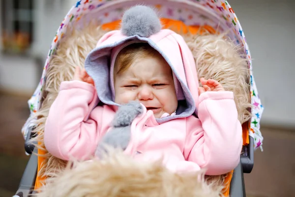 Smutny płacz trochę piękne dziecko dziewczyna siedzi w wózku lub wózka jesienią dnia. Nieszczęśliwy zmęczony i wyczerpany dziecko w ciepłych ubrań, moda stylowy różowy płaszcz dla niemowląt z królika uszy. — Zdjęcie stockowe