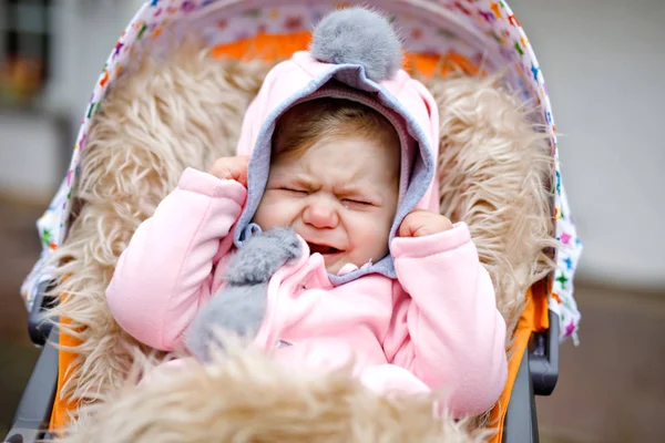 Szomorú sír kis gyönyörű kislány ül a babakocsit vagy babakocsi az őszi napon. Boldogtalan fáradt és kimerült gyermek meleg ruhákat, divat stílusos rózsaszín baba kabát nyuszi füle. — Stock Fotó