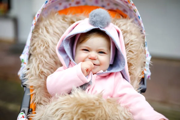 Linda niña hermosa sentada en el cochecito o cochecito en el día de otoño. Feliz niño sonriente en ropa de abrigo, abrigo de bebé rosa con estilo de moda con orejas de conejo. Bebé dando un paseo con los padres. — Foto de Stock