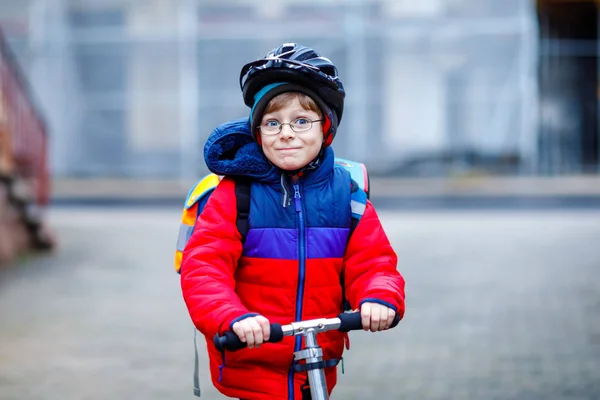 Прекрасный маленький школьник едет на скутере по пути в начальную школу. Ребенок в защитном шлеме, школьная сумка дождливым осенним или зимним холодным утром. Движение в городе и школьники — стоковое фото