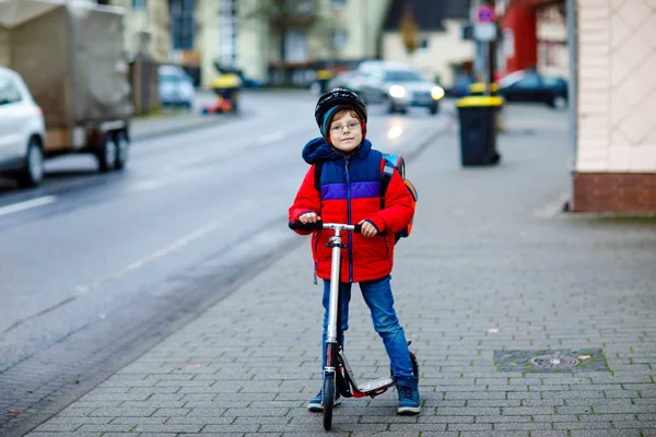 Magnifique petit garçon de l'école chevauchant en scooter sur le chemin de l'école primaire. Enfant avec casque de sécurité, sac d'école le matin froid d'automne ou d'hiver pluvieux. Trafic dans la ville et les écoliers — Photo