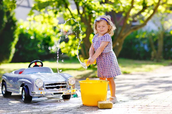 Carino splendida bambina lavare grande vecchia macchina giocattolo nel giardino estivo, all'aperto. Buon bambino sano pulizia auto con acqua e sapone, divertirsi con spruzzi e giocare con la spugna. — Foto Stock