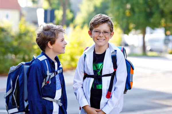 Two little kid boys with backpack or satchel. Schoolkids on the way to school. Healthy adorable children, brothers and best friends outdoors on the street leaving home. Back to school. Happy siblings.