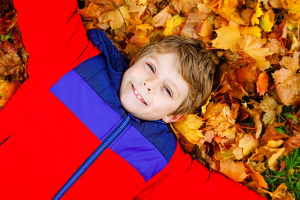 Little kid boy lying in autumn leaves in colorful fashion fall clothing. Happy healthy child having fun in autumn park on warm day. Cute school boy smiling and laughing — Stock Photo, Image
