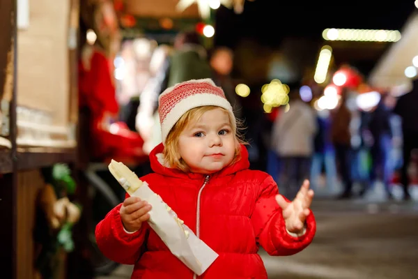 Babymeisje, schattig kind eten van bananen bedekt met chocolade, marshmellows en kleurrijke hagelslag in de buurt van zoete stand met peperkoek en noten. Gelukkig peuter op de kerstmarkt in Duitsland. — Stockfoto