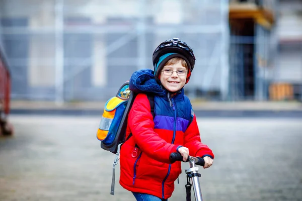 Gorgeous little school kid boy riding on scooter on way to elementary school. Child with safety helmet, school bag on rainy autumn or winter cold morning. Traffic in the city and schoolchildren — Stock Photo, Image