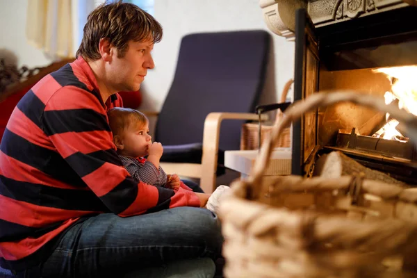 Middle age father sitting by fireplace with cute little baby girl at home. Happy family, dad with daughter make fire for Christmas time. Safe handling for kids