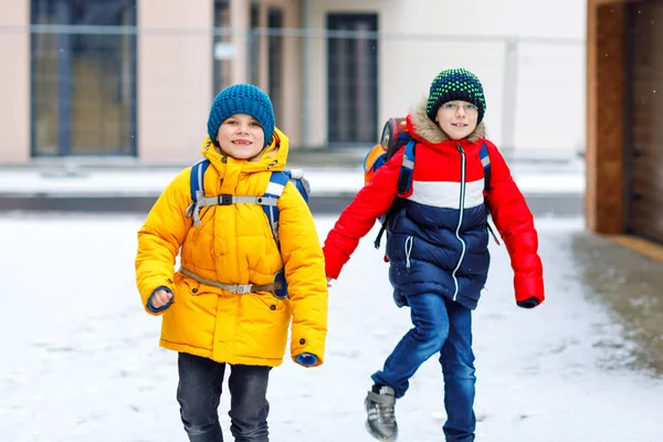 Två små pojkar i grundskolan som går till skolan under snöfallet. Glada barn som har roligt och leker med första snön. Syskon och bästa vän med ryggsäck i färgglada vinterkläder. — Stockfoto