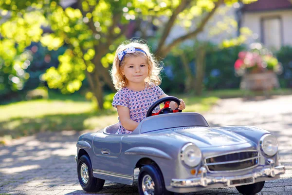 Kis aranyos kisbaba lány vezetés nagy vintage játék autó és szórakozás játszik a szabadban. Gyönyörű, boldog, egészséges gyermek élvezi a meleg nyári napot. Mosolygó lenyűgöző gyerek játszik a hazai kertben — Stock Fotó