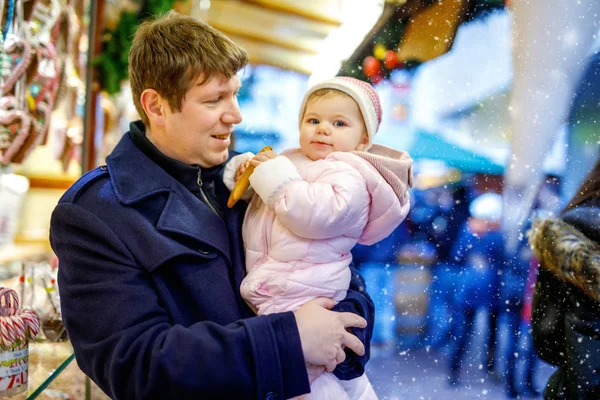 Vader van middelbare leeftijd houdt dochtertje bij zoete stand met peperkoek en noten. Gelukkige familie op kerstmarkt in Duitsland. Schattig meisje dat koekjes eet genaamd Lebkuchen. Feestdag kerstvakantie. — Stockfoto