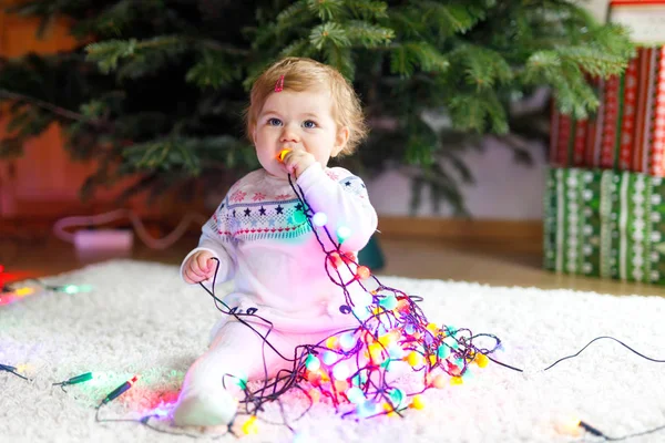 Adorable baby girl holding colorful lights garland in cute hands. Little child in festive clothes decorating Christmas tree — Stock Photo, Image