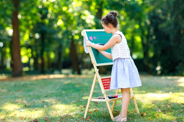 Happy little kid girl standing by big chalk desk Preschool or schoolkid on first day of elementary class. Back to school concept. Healthy child writing and painting on desk outdoors. Copyspace on desk — Stock Photo, Image