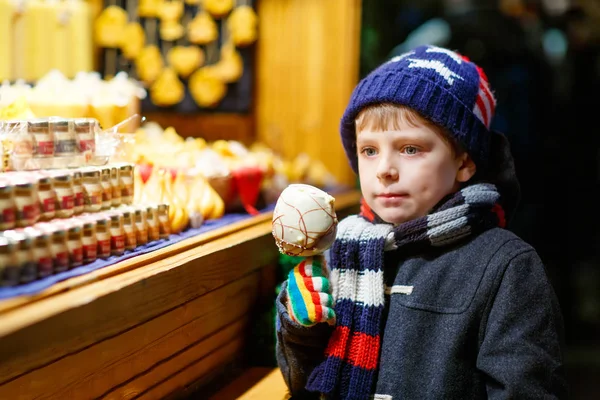 Pequeño niño lindo cerca de soporte dulce con pan de jengibre y nueces. Feliz niño comiendo en manzana cubierta de chocolate blanco. Dulce tradicional en el mercado alemán de Navidad .. —  Fotos de Stock