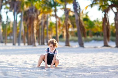 Miami sahilinde eğlenen sevimli, aktif bir çocuk, Key Biscayne. Mutlu şirin çocuk rahatlıyor, kumla oynuyor ve palmiyelerin ve okyanusun yanında güneşli bir günün tadını çıkarıyor.