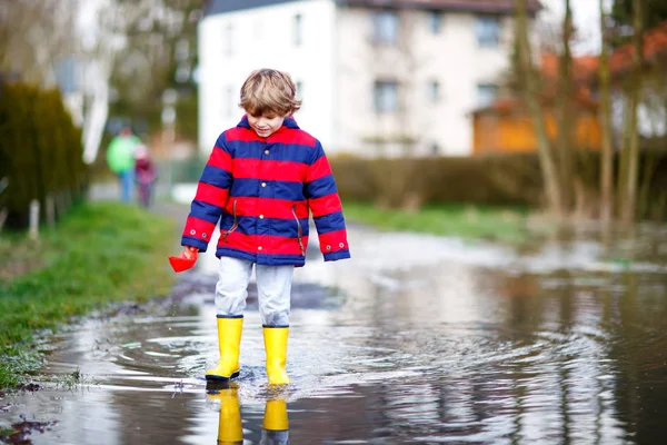 Happy little kid boy in yellow rain boots playing with paper ship boat by huge puddle on spring or autumn day. Active leisure for children. Funny child having fun outdoors, wearing colorful clothes. — Stock Photo, Image