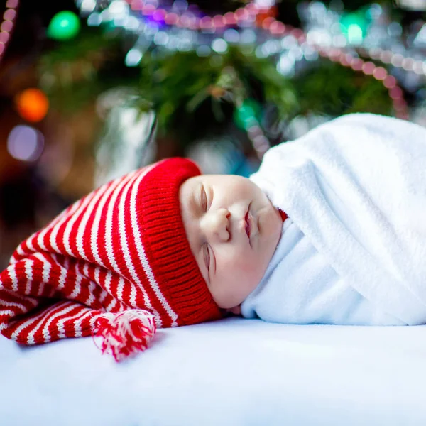 Uma semana de idade bebê recém-nascido em chapéu de Papai Noel envolto em cobertor perto da árvore de Natal com luzes coloridas guirlanda no fundo. Fechar a boca de uma criança gira, um bebé a dormir. Família, Natal, nascimento, nova vida — Fotografia de Stock