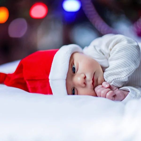Недельный новорожденный ребенок в шляпе Санты, завернутый в одеяло возле елки с красочными гирляндами на заднем плане. Крупный план милого ребенка, спящего маленького ребенка. Семья, Рождество, рождение, новая жизнь — стоковое фото