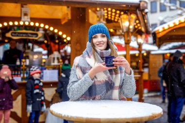 Güzel genç bir kadın sıcak punç içiyor, Alman Noel pazarında sıcak şarap içiyor. Kış kıyafetleri içinde mutlu bir kız. Arka planda ışıklar var. Berlin, Almanya 'da kış günü kar tatilinde..