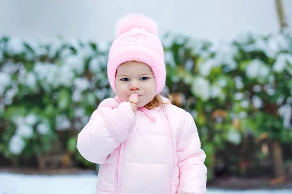 Ritratto di bambina che cammina all'aperto in inverno. Carino il bambino che mangia dolci caramelle lecca lecca. Bambino che si diverte nella fredda giornata sulla neve. Indossare vestiti caldi rosa bambino e cappello con ciambelle. — Foto Stock