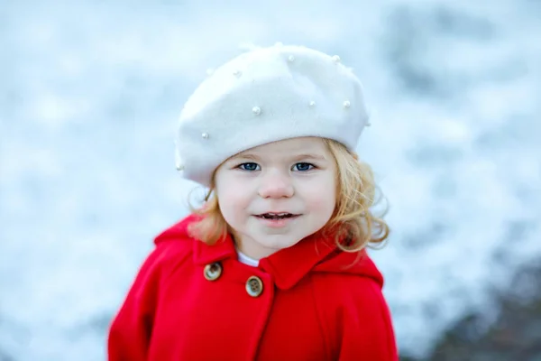 Outdoor winter portrait of little cute toddler girl in red coat and white fashion hat barret. Healthy happy baby child walking in the park on cold day with snow and snowfall. Stylish clothes for kids. — Stock Photo, Image