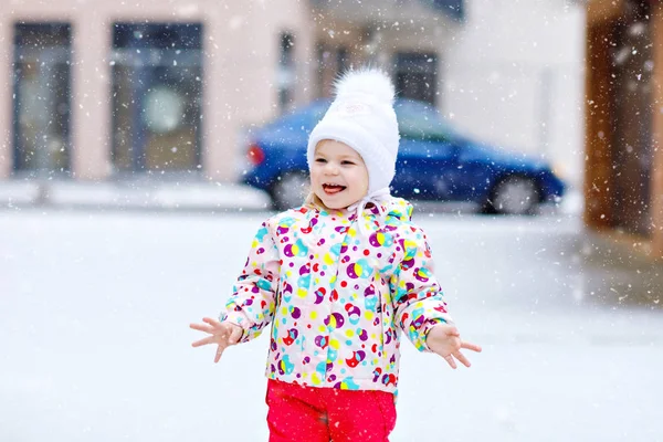 Portret van een klein peutermeisje dat 's winters buiten loopt. Leuke peuter die zoete lolly snoep eet. Kind heeft plezier op koude sneeuwdag. Het dragen van warme baby kleurrijke kleding en hoed met bobbels. — Stockfoto