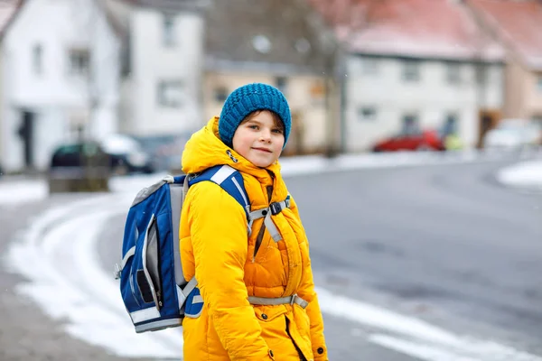 Niño de la escuela de primaria caminando a la escuela durante las nevadas. Niño feliz divirtiéndose y jugando con la primera nieve. Estudiante con chaqueta amarilla y mochila en ropa de invierno colorida . — Foto de Stock