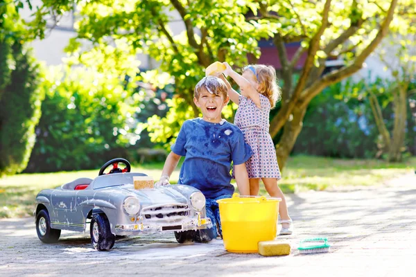 Two happy children washing big old toy car in summer garden, outdoors. Brother boy and little sister toddler girl cleaning car with soap and water, having fun with splashing and playing with sponge. — Stock Photo, Image
