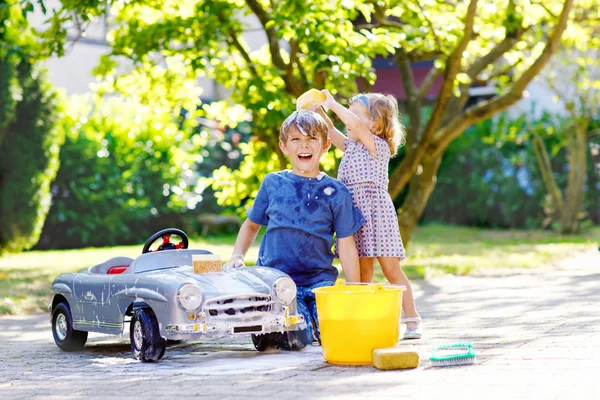 Duas crianças felizes lavando grande carro de brinquedo velho no jardim de verão, ao ar livre. Irmão menino e irmã mais nova menina de limpeza carro com sabão e água, se divertindo com espirrar e brincar com esponja. — Fotografia de Stock