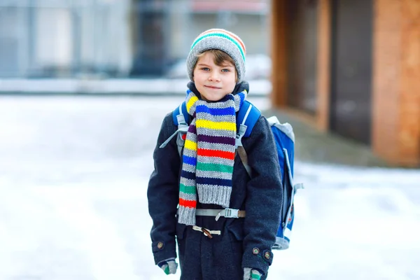 En liten skolpojke i grundskolan som går till skolan under snöfallet. Glada barn som har roligt och leker med första snön. Studerande med ryggsäck eller väska i färgglada vinterkläder. — Stockfoto