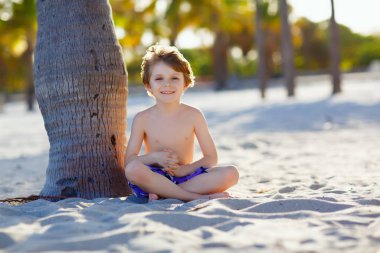 Sarışın, tapılası, yakışıklı çocuk Miami sahilinde eğleniyor, Key Biscayne. Kumla oynayan ve okyanus kenarında koşan sağlıklı, mutlu bir çocuk. Palmiyeler, güvenlik evi ve beyaz kum.