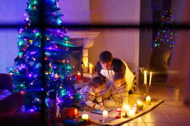 Baba ve Noel arifesi zamanında şömine baca tarafından oturan iki küçük çocuğu. Baba ve çocuklar çocuklar oğulları tablet ile oynuyor. Açık havada kar yağışı sırasında gelen pencereden. Aile xmas kutluyor.