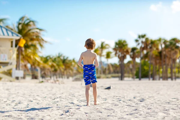 Niño rubio divirtiéndose en la playa de Miami, Key Biscayne. Feliz niño lindo saludable jugando con arena y corriendo cerca del océano. Palmeras, casa de seguridad y arena blanca — Foto de Stock