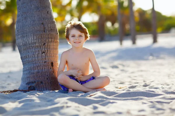 Rubio adorable niño precioso divirtiéndose en la playa de Miami, Key Biscayne. Feliz niño lindo saludable jugando con arena y corriendo cerca del océano. Palmeras, casa de seguridad y arena blanca — Foto de Stock