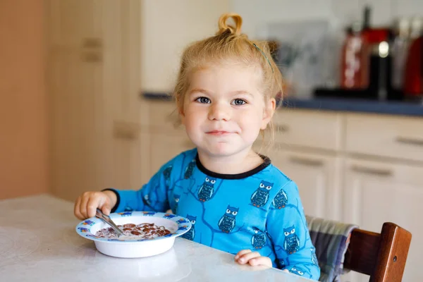 Linda menina criança comendo cereais saudáveis com leite para o café da manhã. Bonito bebê feliz criança em roupas coloridas sentado na cozinha e se divertindo com a preparação de aveia, cereais. Interior em casa — Fotografia de Stock