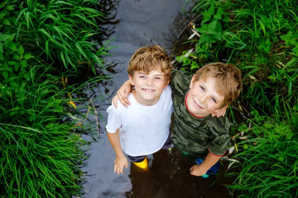 Deux garçons heureux petits écoliers, drôles de frères et sœurs s'amusant ensemble à marcher dans l'eau dans la rivière dans des bottes en gomme de caoutchouc. Portrait de famille de frères et de meilleurs amis en bonne santé — Photo