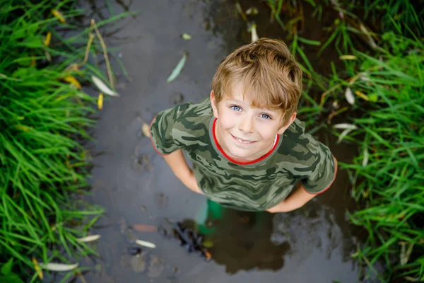 Ευτυχισμένο μικρό σχολείο παιδί που διασκεδάζει περπατώντας μέσα από το νερό στο ποτάμι με μπότες από λαστιχένια τσίχλα. Παιδικό πορτρέτο του υγιούς παιδιού στις καλοκαιρινές διακοπές, ενεργά παιχνίδια σε εξωτερικούς χώρους — Φωτογραφία Αρχείου
