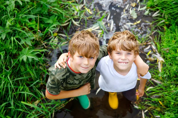 Deux garçons heureux petits écoliers, drôles de frères et sœurs s'amusant ensemble à marcher dans l'eau dans la rivière dans des bottes en gomme de caoutchouc. Portrait de famille de frères et de meilleurs amis en bonne santé — Photo